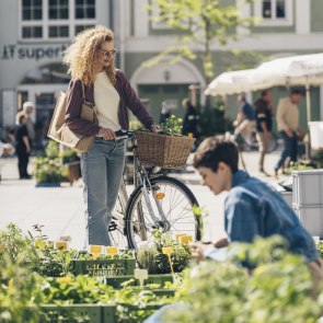 Frau mit Rad am Markt am Domplatz sowie weitere Frau kniet bei einem Stand und begutachtet Kräuter